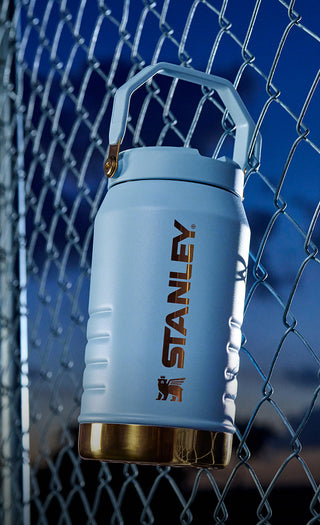 Light blue Stanley insulated bottle with gold base against a chain-link fence.