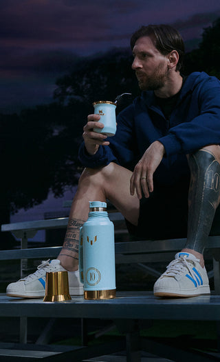 Messi sitting outdoors at night holding a drink container with a blue water bottle and gold cups on a table.