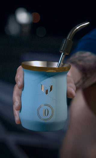 Hand holding a blue mug with a gold lid and straw, blurred background
