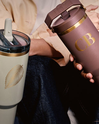 Two people holding insulated travel mugs, one with a leaf design and the other with 'CB' branding.