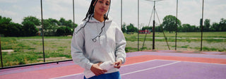 Person standing on a purple tennis court wearing a gray hoodie and blue shorts.