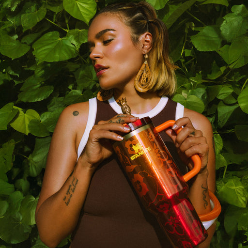 Karol G holding a red Stanley tumbler against a green leafy background