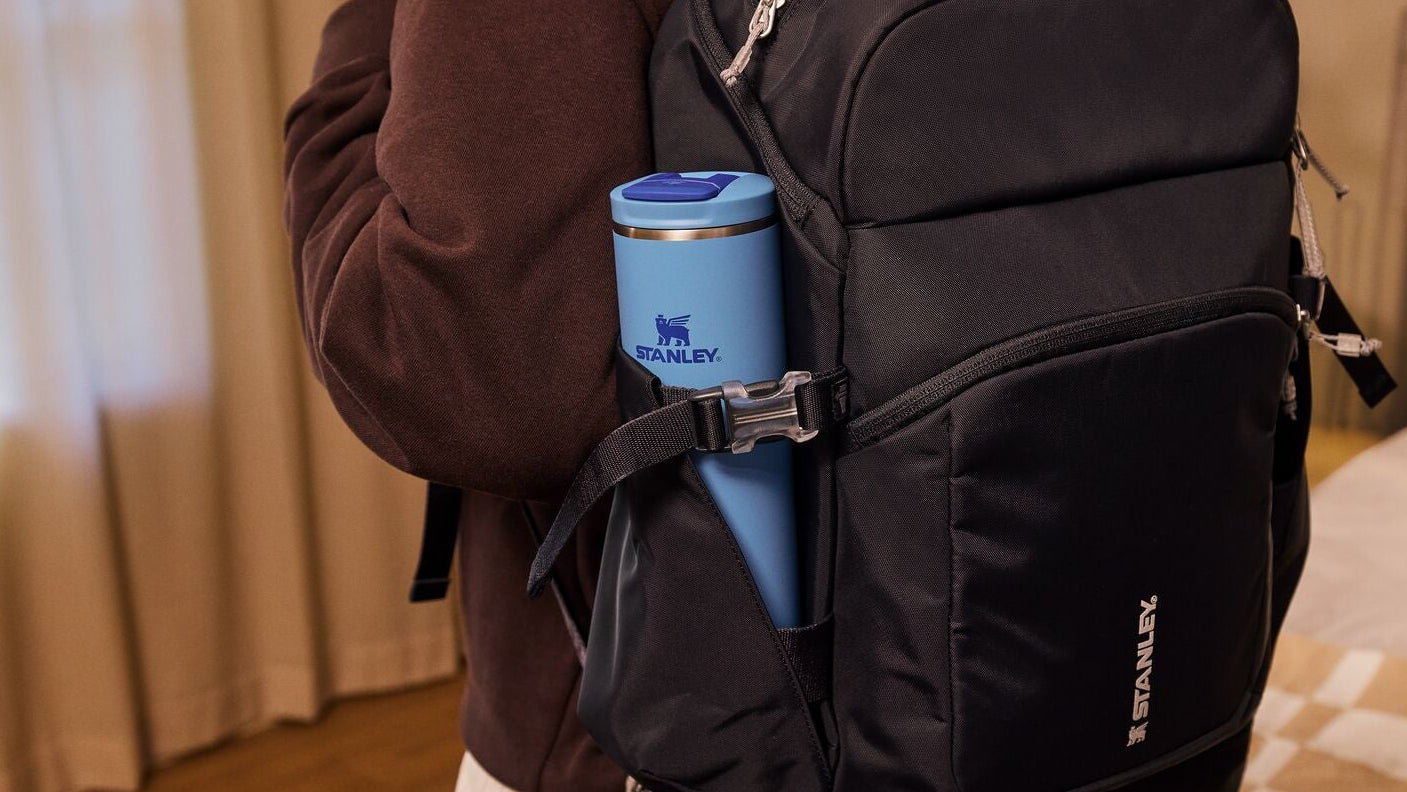 Person wearing a black backpack with a blue thermos in a room setting.