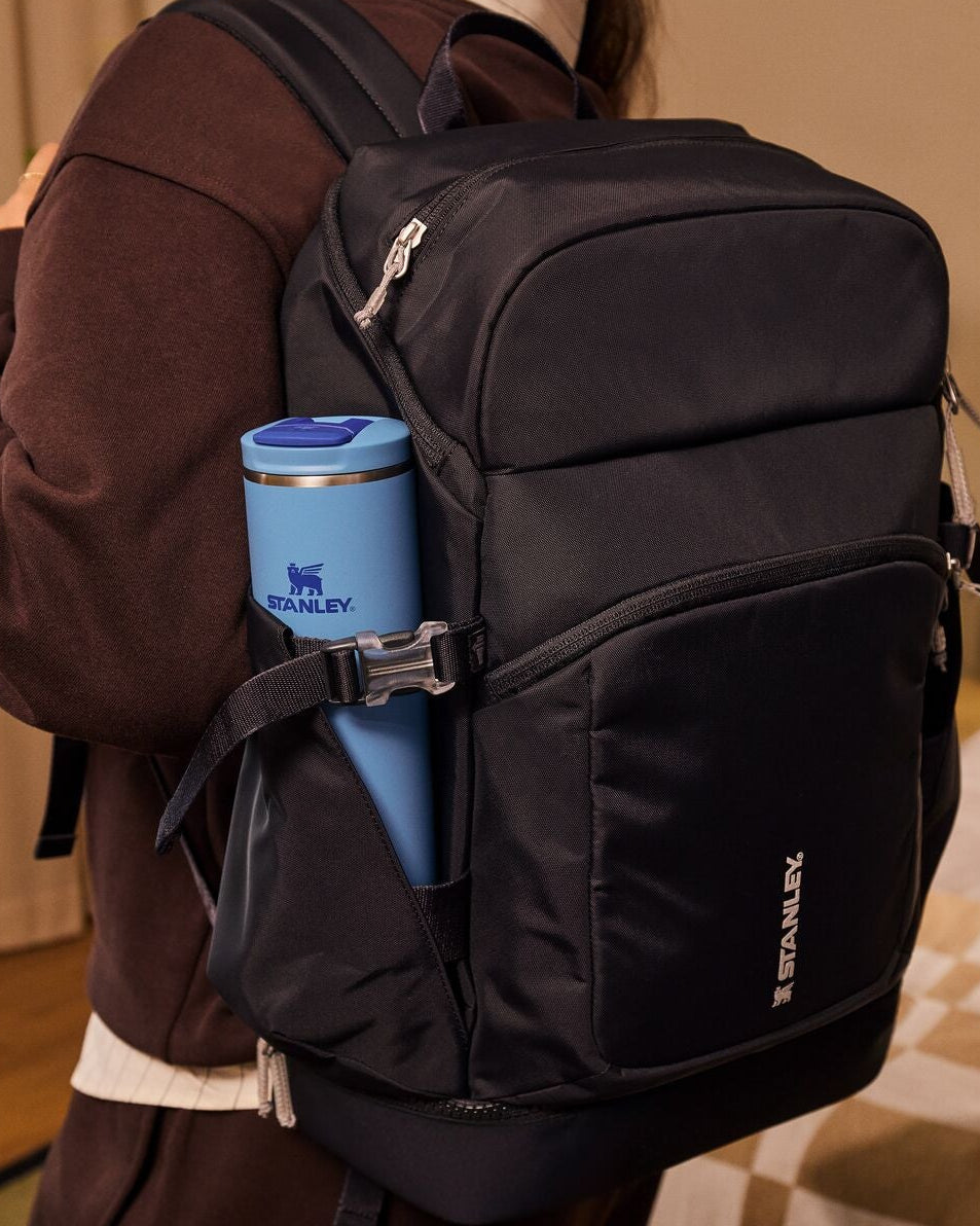 Person wearing a black backpack with a blue thermos in a room setting.