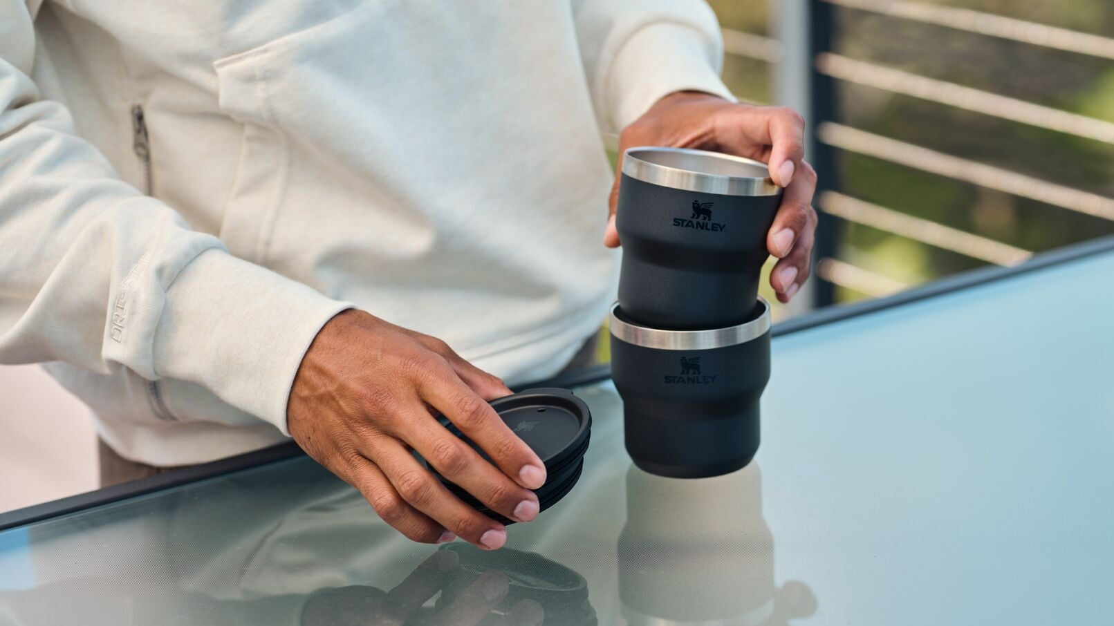 Person holding a black stacking mug on a reflective surface with a blurred outdoor background