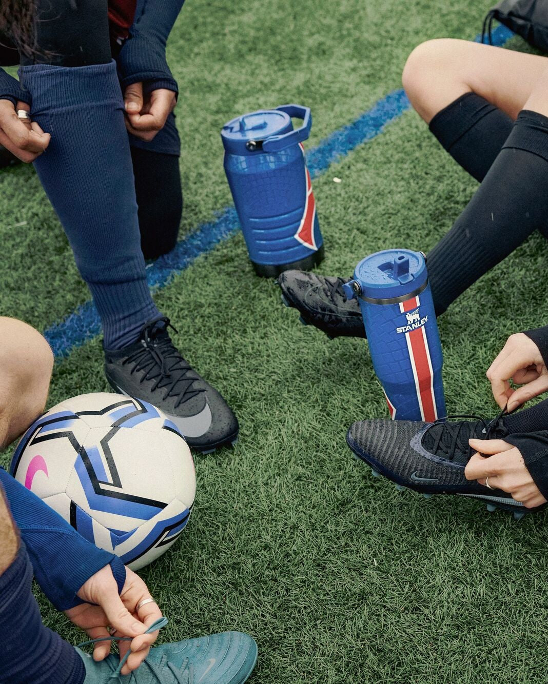 People sitting on a grassy field with soccer balls and water bottles.