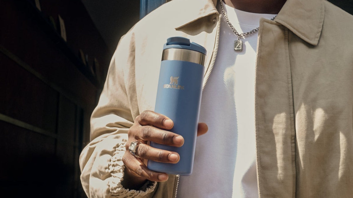 Man holding a blue Stanley travel mug in front of a coffee place.